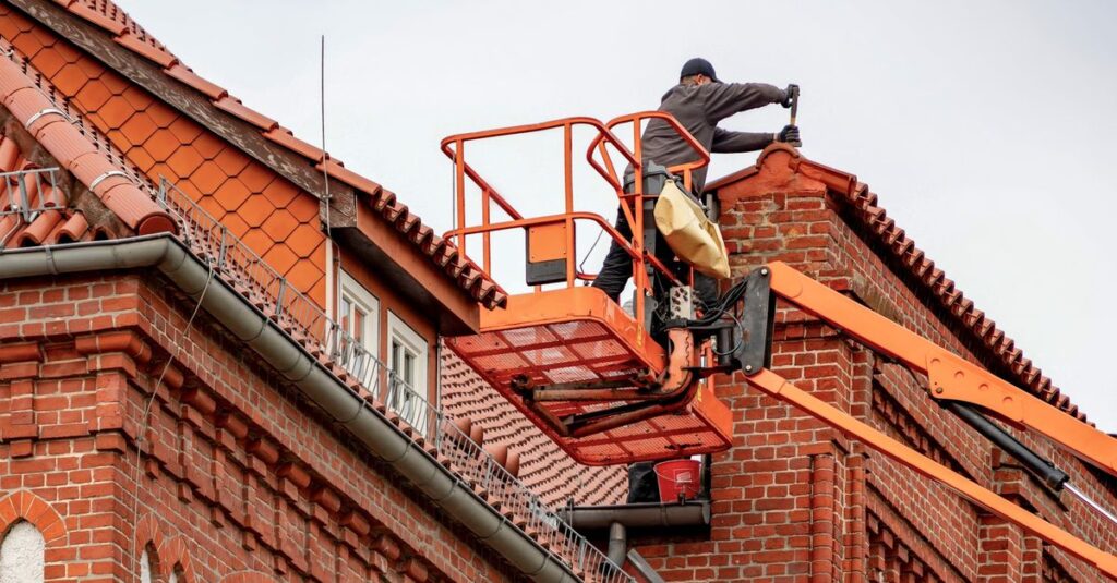 Roofer in a cherry picker conducting maintenance on a red brick building with a tiled roof.