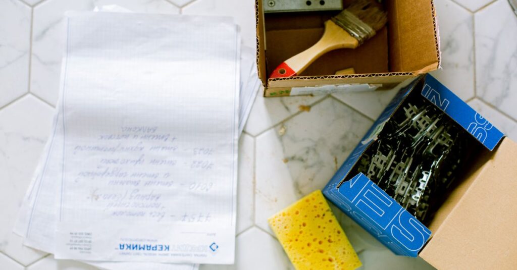 Flat lay of home renovation tools including paintbrush, sponge, and hardware on tiled floor.