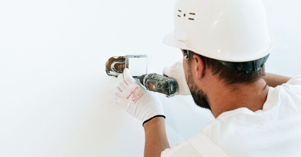 Construction worker wearing helmet and gloves uses a power tool to fix a wall.