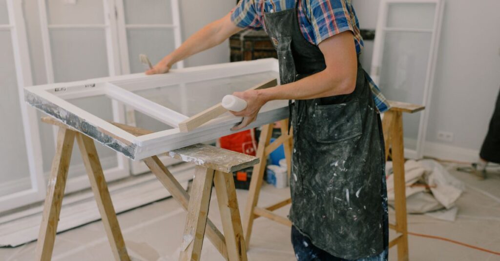 Unrecognizable male master wearing dirty apron standing near window with white frames on wooden workbench while working in professional studio