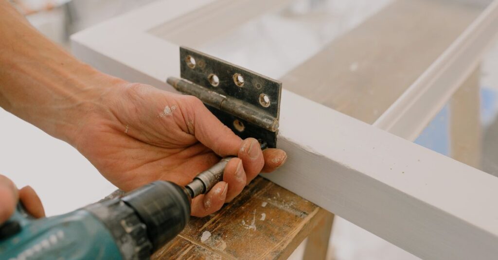 Crop unrecognizable worker drilling metal hinge on white window frame during house renovation