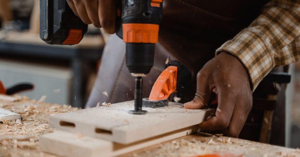 Carpenter uses drill on wood board, surrounded by sawdust in a workshop setting.