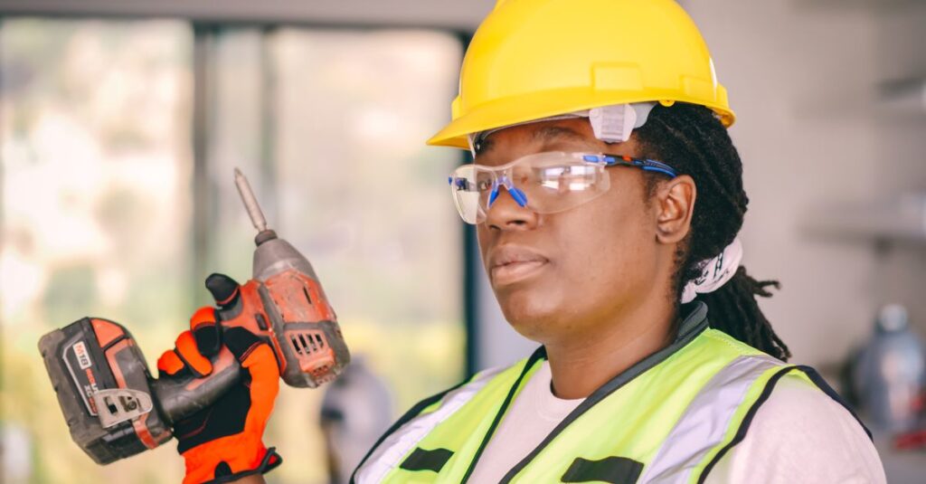 Confident female construction worker wearing safety gear and holding a drill indoors.